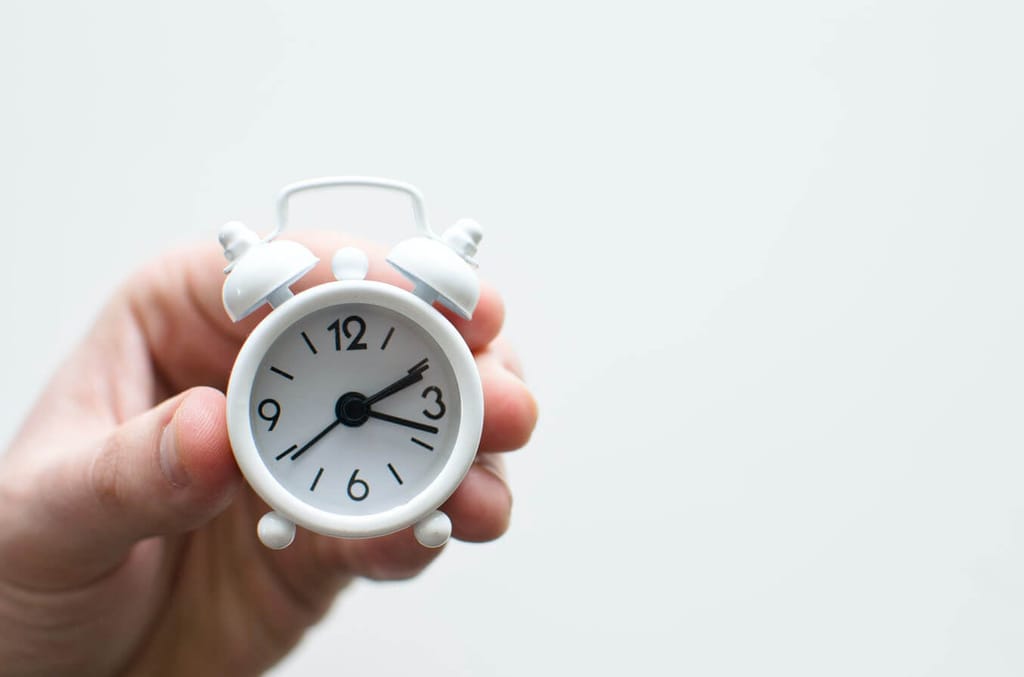 A hand holding a small, white, analog alarm clock.