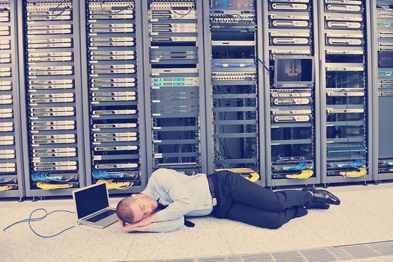 A man sleeping on the floor next to his lap top, in front a wall of servers.