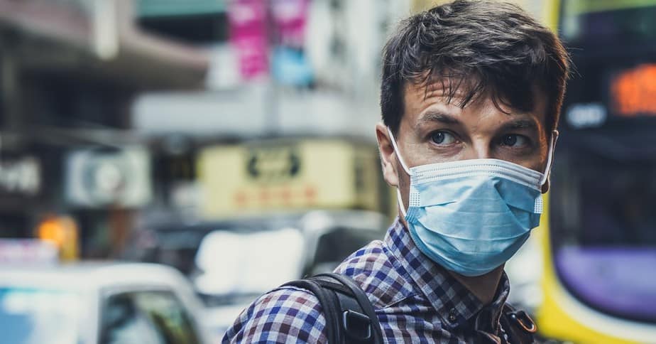 A man wearing a surgical mask and a plaid shirt, in front of city traffic.