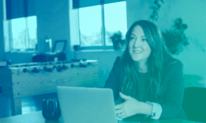 A woman sitting at a table with a laptop, looking up as if listening to someone else.