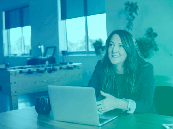 A woman sitting at a table with a laptop, looking up as if listening to someone else.