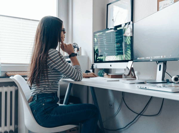 A woman in a home office sitting at a white desk with 2 monitors, typing on a keyboard, on a computer with productivty mangement.