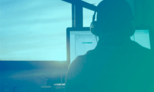 Seen from behind, a man wearing headphones and working on his computer. He is facing a sunrise.
