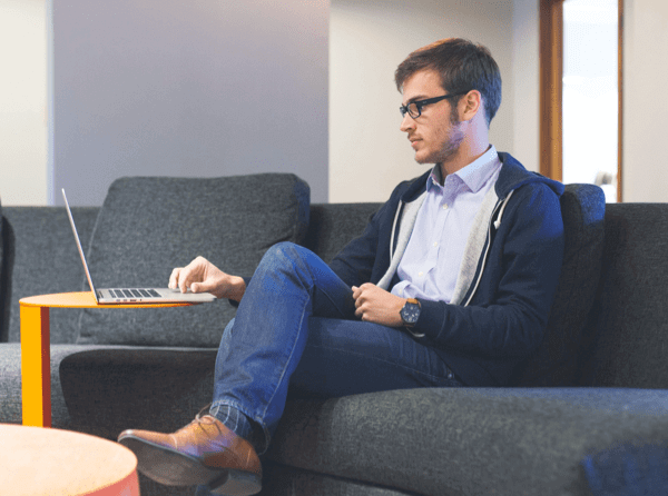 A man wearing a jacket, jeans and brown shoes, sitting on a couch in a waiting room while working on his laptop.