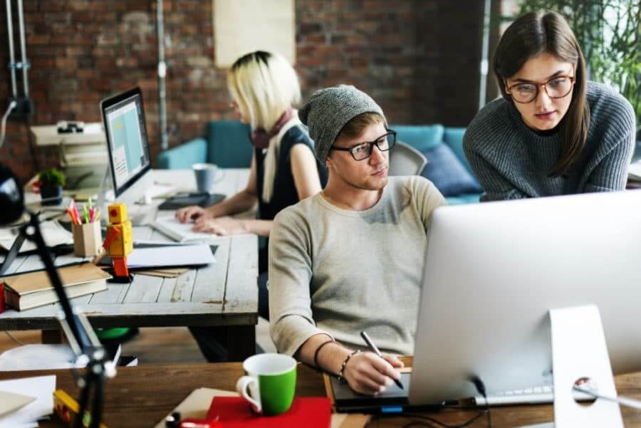 A woman in the background working at a top on a computer, a man at a desk looking at a monitor with a woman next to him.