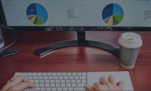 Hands on a wireless keyboard in front of a computer monitor showing charts and graphs. A coffee cup is to the right.