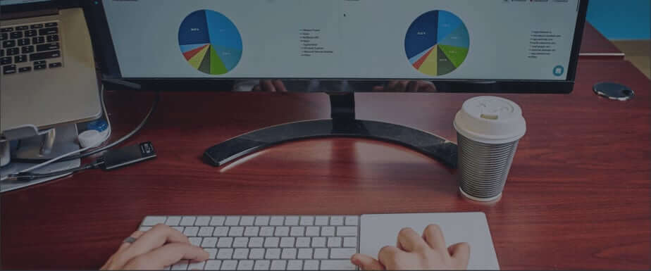 Hands on a wireless keyboard in front of a computer monitor showing charts and graphs. A coffee cup is to the right.