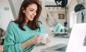 A woman in teal sweater holds a coffee much while looking at her laptop while working from home.