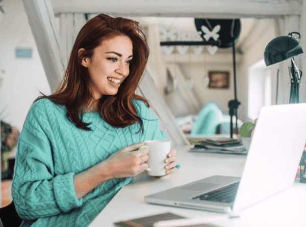 A woman in teal sweater holds a coffee much while looking at her laptop while working from home.