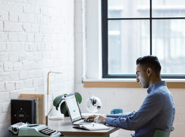 A man in a blue checkered shirt sits at a desk working from home on his laptop, and a typewriter to his left.