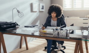 A woman working from home, at a glass top desk. She's writing with a pen and has a printer and lamp on the desk.