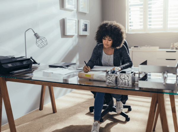 A woman working from home, at a glass top desk. She's writing with a pen and has a printer and lamp on the desk.