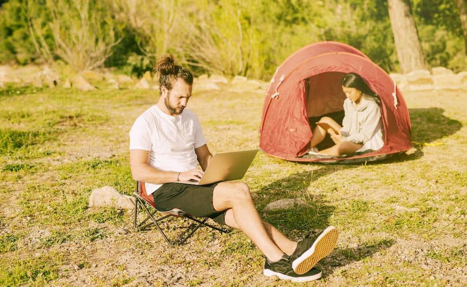 A man sitting in a small camping chair working on his laptop and a woman inside a red tent, work on her tablet.