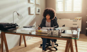 A woman working in a home office at a glass top desk. She's writing with a pen and has a printer and lamp on the desk.