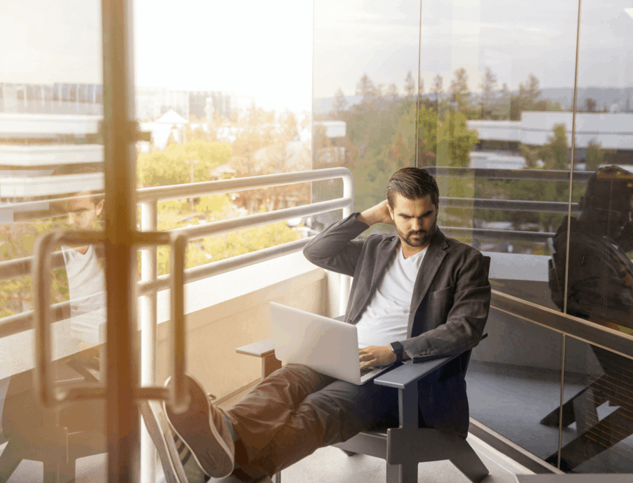 A man sitting on a balcony working on his laptop, which runs productivity monitoring software, on an upper story.