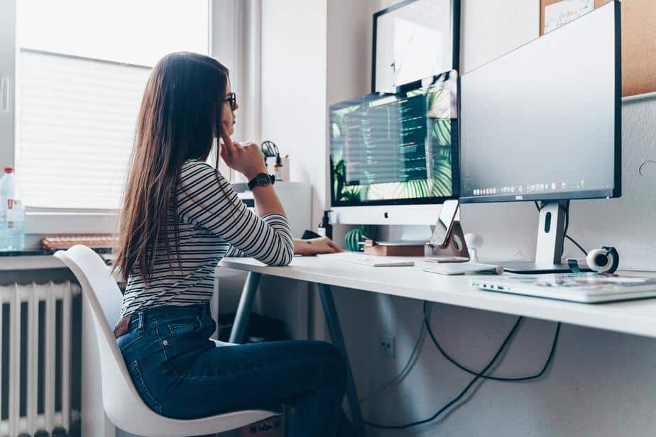 A woman in a home office sitting at a white desk with 2 monitors, typing on a keyboard, on a computer productivity management.