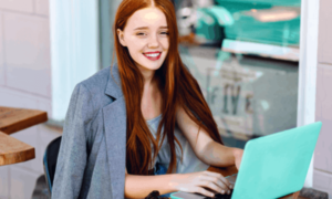 A girl with long red hair sits out an outdoor table while working on a sea green laptop with productivity measurement software.