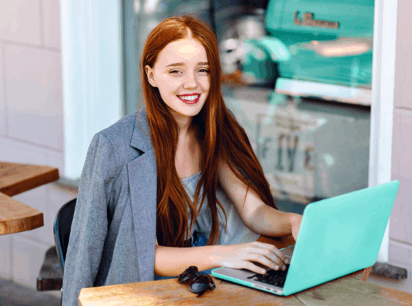 A girl with long red hair sits out an outdoor table while working on a sea green laptop with productivity measurement software.