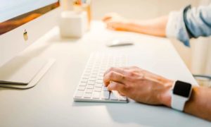 Hands typing on a keyboard of an Apple computer, which uses productivity measurement software and wearing an apple watch.