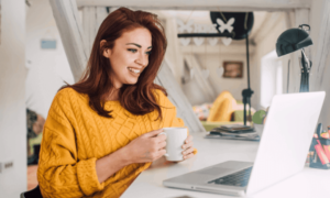 A woman in gold sweater holds a coffee much while looking at her laptop while working from home.