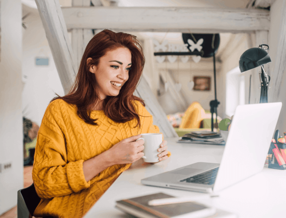 A woman in gold sweater holds a coffee much while looking at her laptop while working from home.