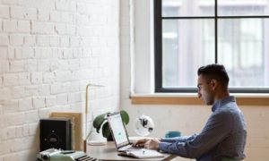A man in a blue checkered shirt sits at a desk working from home on his laptop, and a typewriter to his left.
