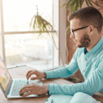 A man in a mint green shirt is working on his laptop, with remote workforce management software, in his home office.