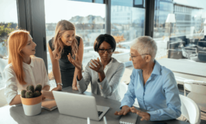 4 women gathered around a table in an office, which has a laptop, notebooks and a small cactus plant on it.