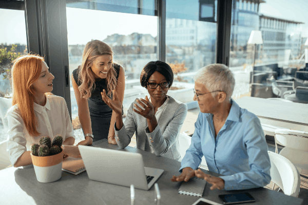 4 women gathered around a table in an office, which has a laptop, notebooks and a small cactus plant on it.