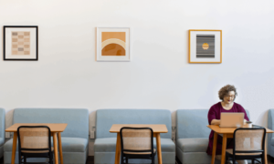 A woman working on her laptop, which uses productivity measurement software, in a café with small, blue couches.