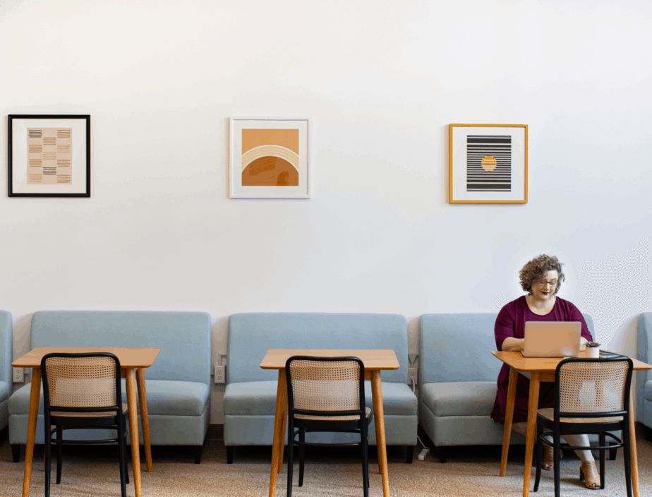 A woman working on her laptop, which uses productivity measurement software, in a café with small, blue couches.