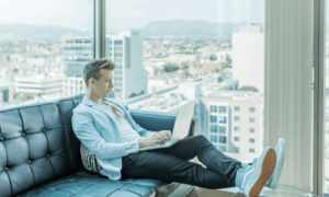 A man sitting on a couch, working on his laptop, with productivity monitoring, on a high level with a view overlooking a city.