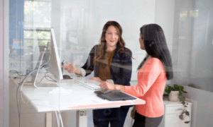 2 women standing at a desk talking. One woman is pointing to the computer monitor showing productivity monitoring software.