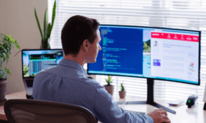 A man working in his home office. He has a large, curved monitor and a laptop with remote workforce management software.