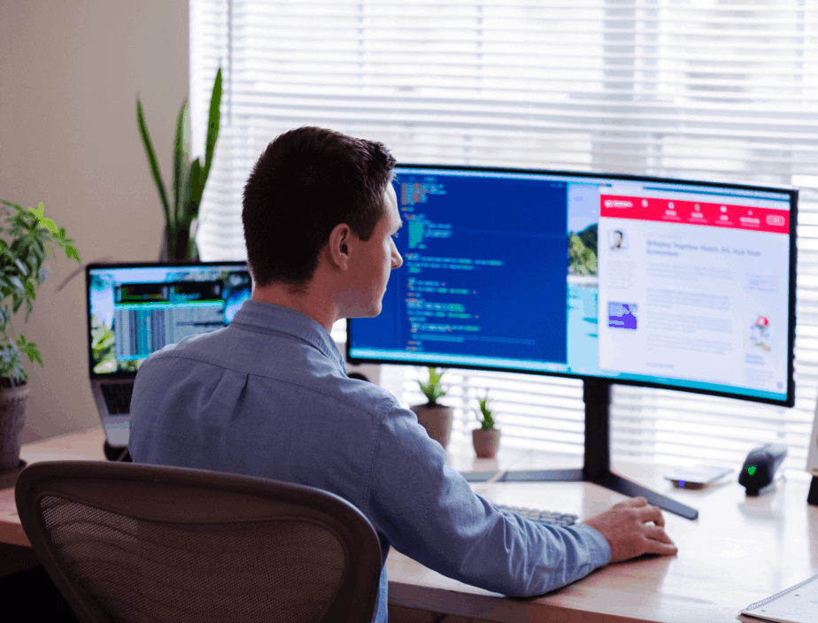 A man working in his home office. He has a large, curved monitor and a laptop with remote workforce management software.