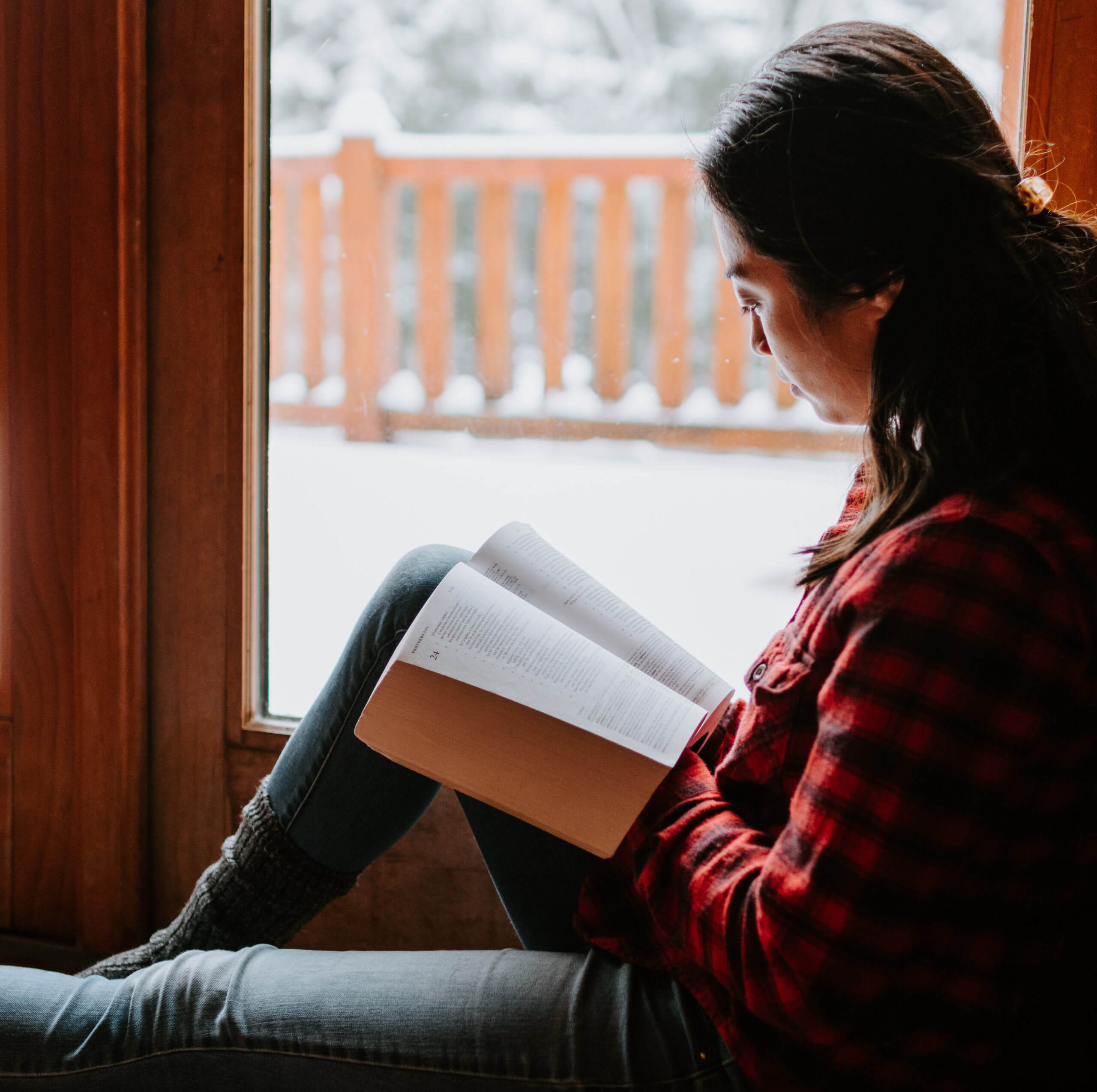 A woman sitting on the floor next to a window, reading a book.