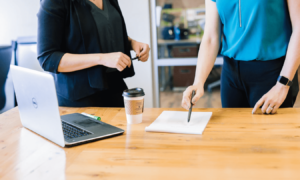 2 women standing at a conference table. One holds a pin with both hands, the other uses a pen to point to words on a notepad.