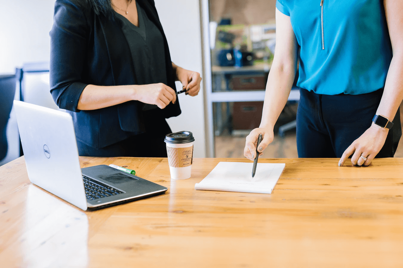 2 women standing at a conference table. One holds a pin with both hands, the other uses a pen to point to words on a notepad.