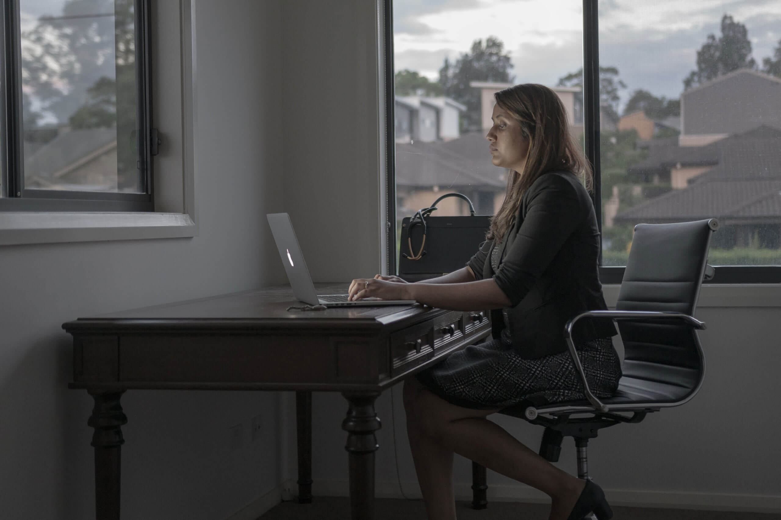 A woman sitting at a large desk in her home office typing on her laptop, which uses remote workforce management software.
