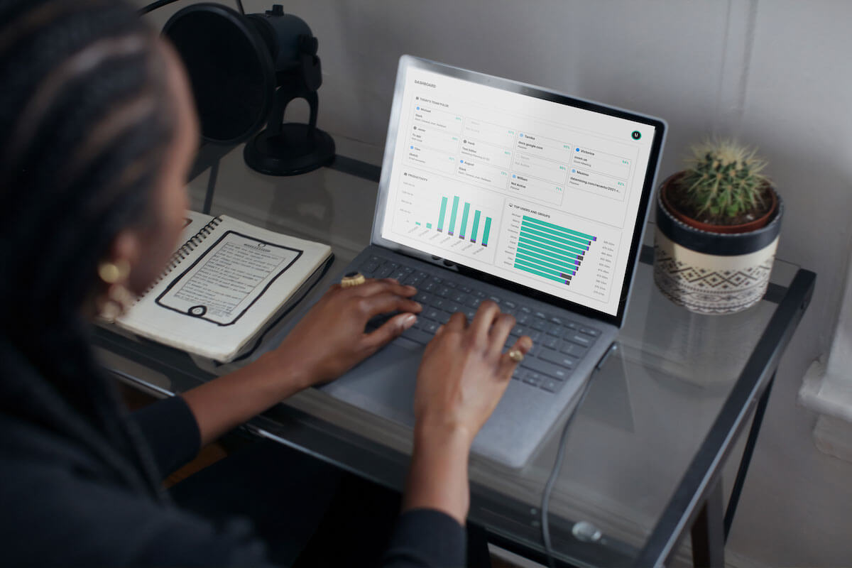 A woman typing on her laptop, which shows and ActivTrak productivity management software, at a small, glass top desk.