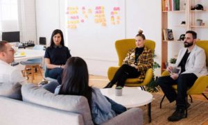 5 people sitting in chairs or on a couch in an office listening to once person speaking. There are post it notes on the wall.
