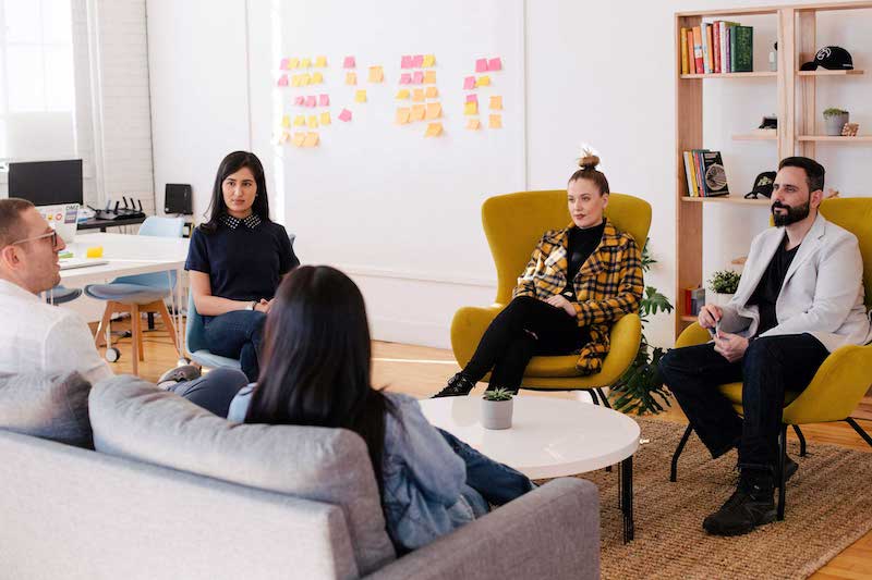 5 people sitting in chairs or on a couch in an office listening to once person speaking. There are post it notes on the wall.