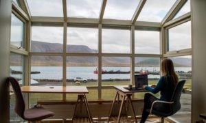 A woman working from home at a small desk with a wall of windows in front of her, looking out on a lake and hills.