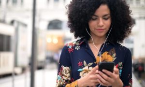 A woman standing on a sidewalk in a city, looking at workforce analytics on her phone with ear buds in her ears.