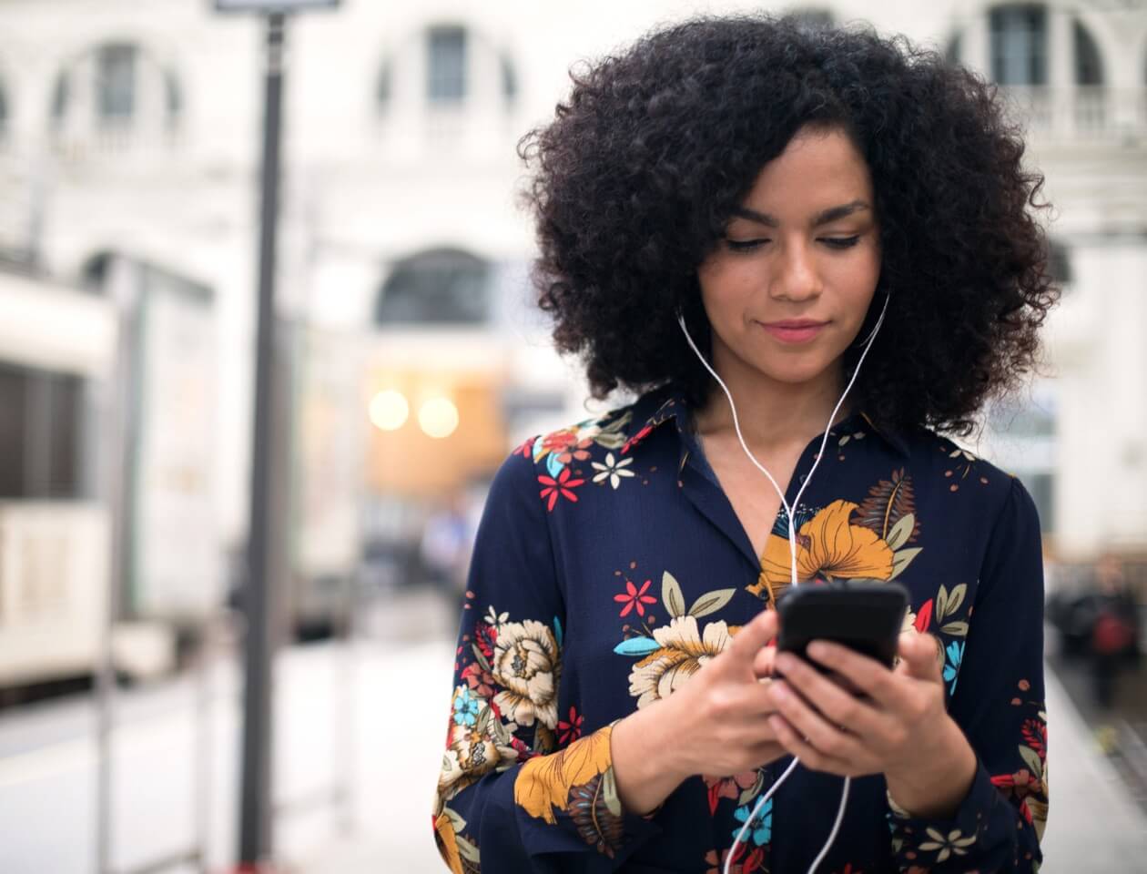 A woman standing on a sidewalk in a city, looking at workforce analytics on her phone with ear buds in her ears.