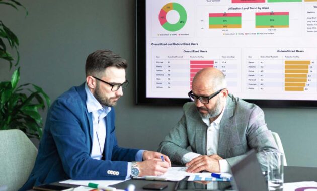 Two people reviewing documents in a meeting room with a data dashboard in the background.