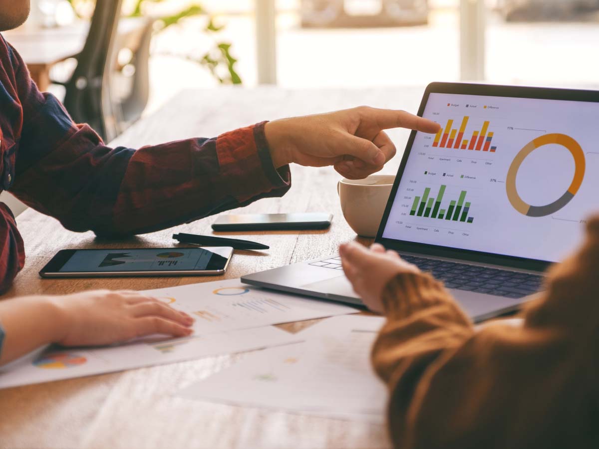Two people discuss colorful charts on a laptop screen at a wooden table, indicating data analysis. Nearby, papers and a tablet are seen, suggesting teamwork.