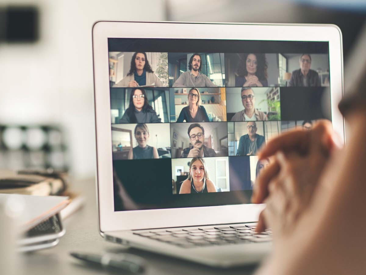 Laptop screen displaying a video conference with twelve diverse people in individual squares. A person’s hands are clasped in the foreground, suggesting attentiveness.