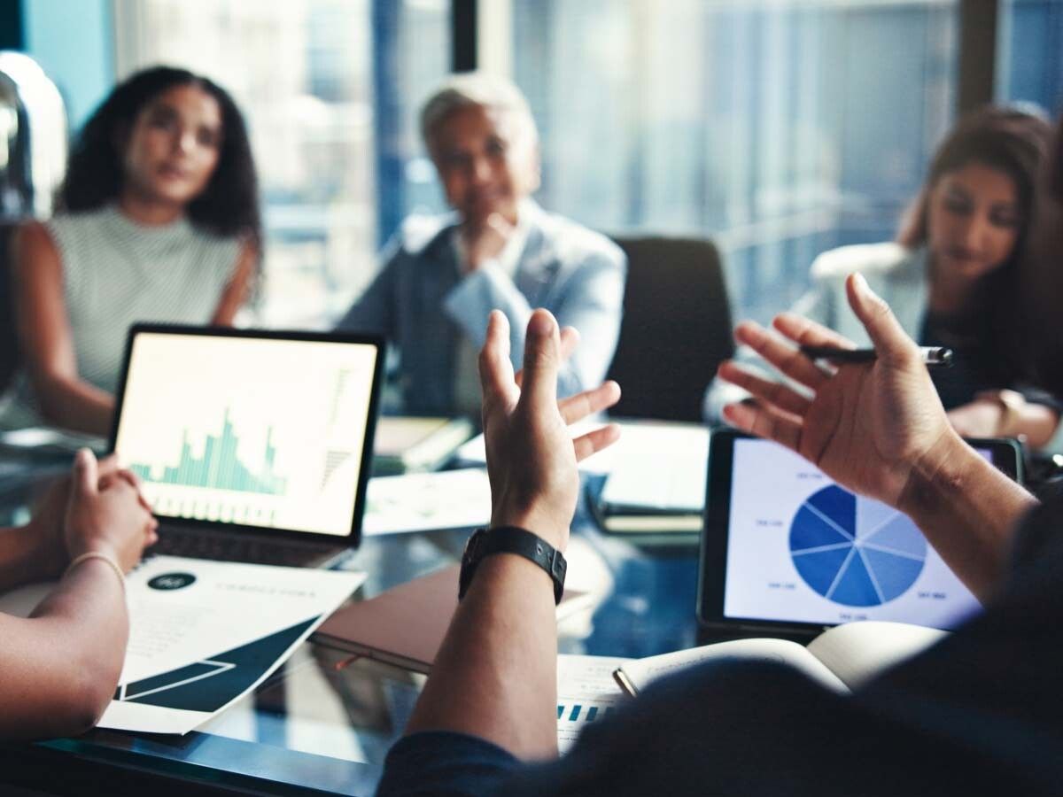 A business meeting with diverse people around a table, papers, and laptops displaying charts and graphs. The focus is on a person gesturing animatedly.