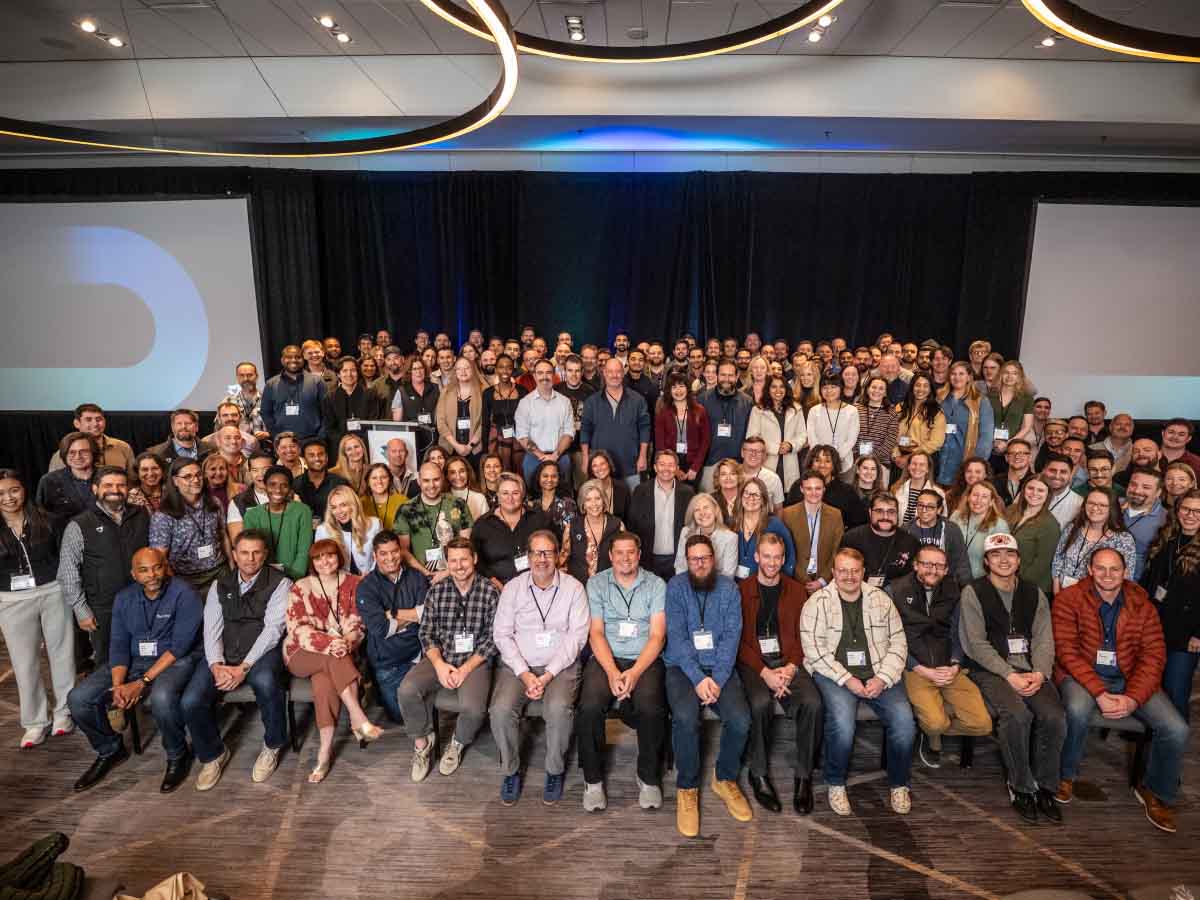 A large group photo of diverse adults in a conference setting. They are smiling, seated and standing in rows on a carpeted floor, with a stage and screens behind them.